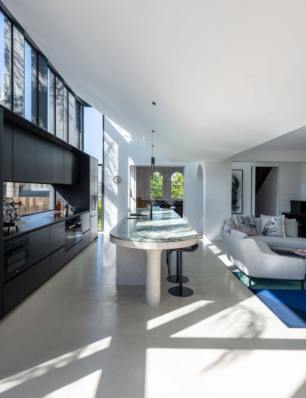 Bright open-plan contemporary interior combining a sleek black kitchen with full-height glazing, a sculptural stone island, and an adjoining living area. White textured walls, polished light flooring, arched openings, and abundant natural light create a refined, airy space.