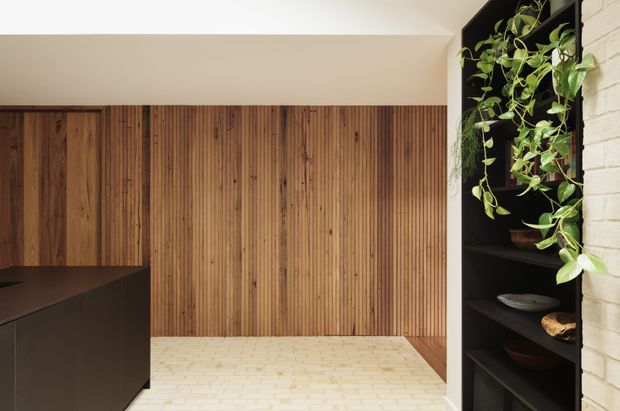 A refined interior corner featuring a warm vertical timber-lined wall, pale brick flooring, dark matte built-in cabinetry, and an open black shelving unit with trailing greenery and ceramics. The space appears to be a contemporary kitchen or adjoining living area, with a partial dark island visible at left and a calm, material-focused composition.