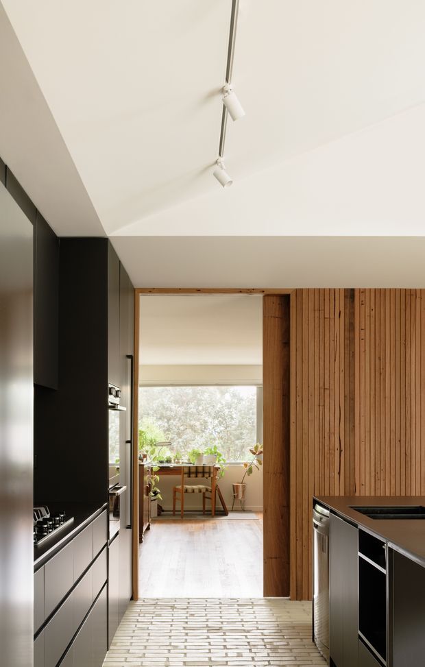 A narrow, contemporary kitchen viewed toward a bright dining or sunroom area. The kitchen features matte dark cabinetry, integrated appliances, a dark benchtop, a central island, and a striking fixed timber slat wall and doorway surround. Pale brick-like flooring leads through the space, while a linear track light runs across the white ceiling. Plants and a small dining table are visible in the sunlit adjoining room.
