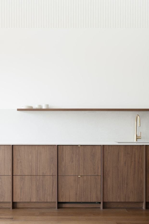 Minimalist kitchen joinery with warm walnut-toned cabinetry, a pale stone or solid-surface benchtop, slim timber wall shelf, and a brass gooseneck tap. The space has a largely white palette, clean horizontal lines, integrated cabinetry, and subtle vertical wall detailing near the ceiling.