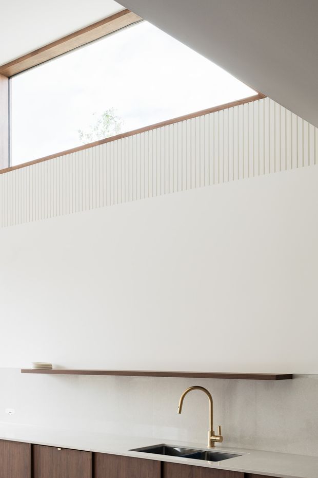 A bright minimalist kitchen interior with dark timber lower cabinetry, a pale stone benchtop and matching backsplash, an undermount stainless-steel sink, and a brass gooseneck tap. A long timber shelf runs across the wall, while a large skylight or clerestory opening with timber framing brings in abundant natural light above vertically ribbed white wall panelling.