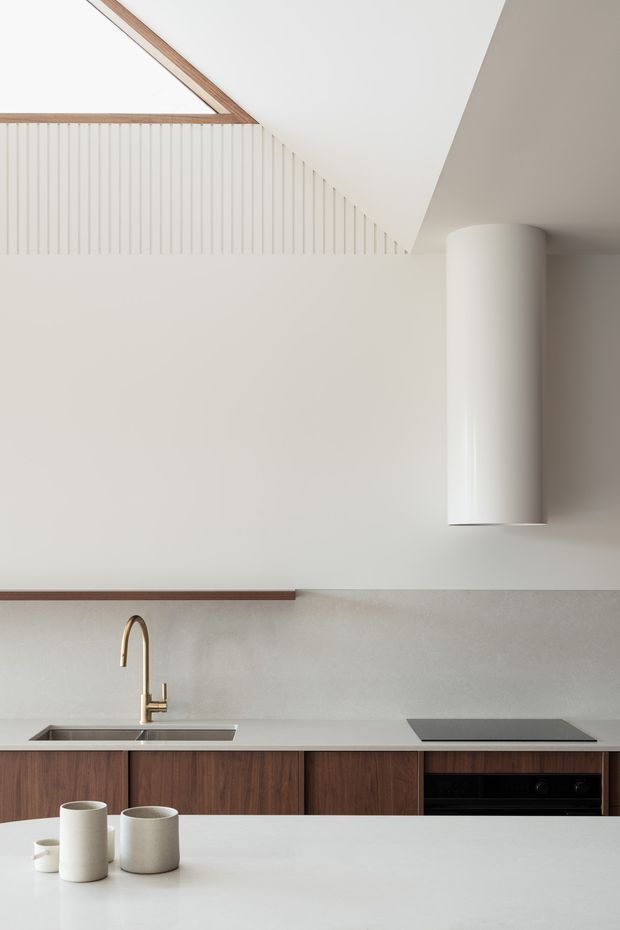 Minimalist contemporary kitchen interior with warm timber cabinetry, a pale stone or composite benchtop and splashback, integrated black cooktop and oven, stainless-steel sink, and a brushed brass gooseneck tap. A large cylindrical white rangehood and vertical fluted wall detailing create strong architectural features, while a skylight or angled ceiling opening introduces natural light.