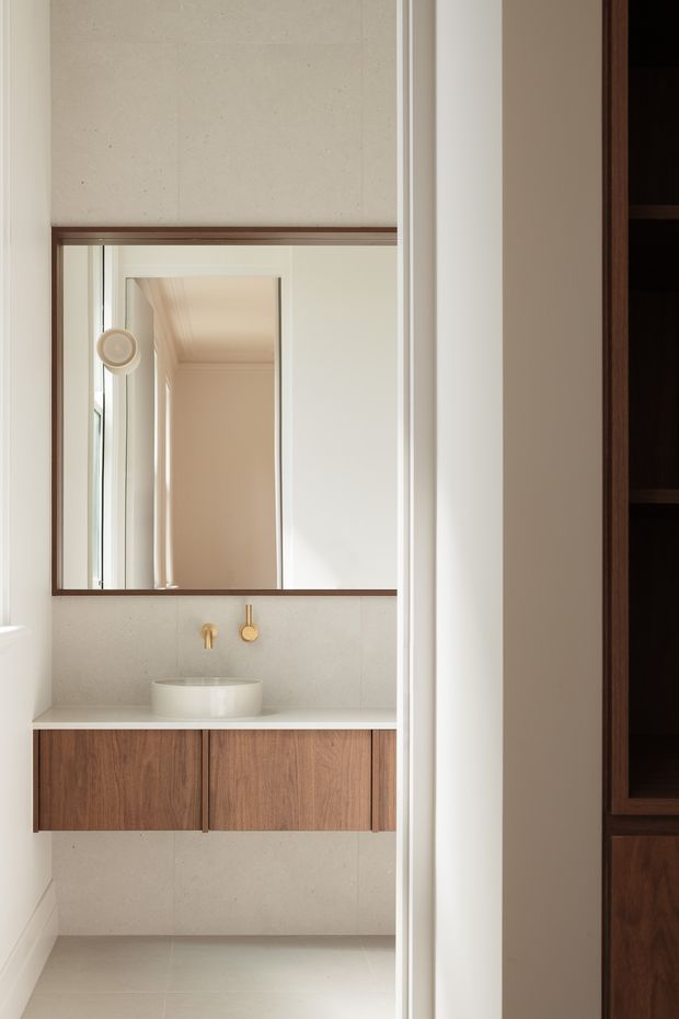 A refined minimalist bathroom vanity viewed through a doorway. The room features a floating warm timber vanity with flat-front drawers, a white vessel basin, a pale stone or concrete-look benchtop and backsplash, warm brass wall-mounted tapware, light tiled flooring, and a large framed mirror. A dark timber built-in cabinet is partially visible at the right edge, while the mirror reflects a softly lit hallway or adjoining space.