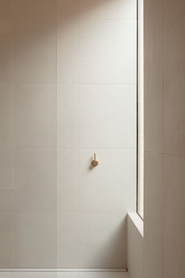 Minimalist close-up of a tiled bathroom or shower enclosure, featuring large-format pale beige and off-white wall tiles, fine grout lines, a small brass-gold control or hook, and a narrow vertical window revealing strong natural light and geometric shadow. The image is architectural and detail-focused, with no visible furniture or broader room context.
