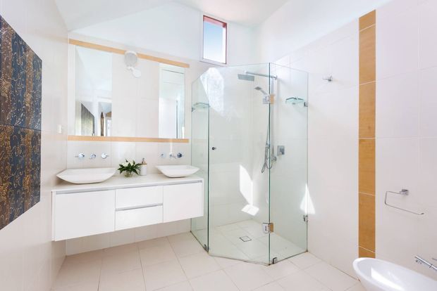 Bright contemporary bathroom with a floating white double vanity, two vessel basins, large wall mirror, frameless glass shower enclosure, wall-mounted fixtures, and pale tiled surfaces. A dark patterned feature tile wall and vertical timber-toned accent strip add contrast. A freestanding bathtub is partially visible in the foreground.