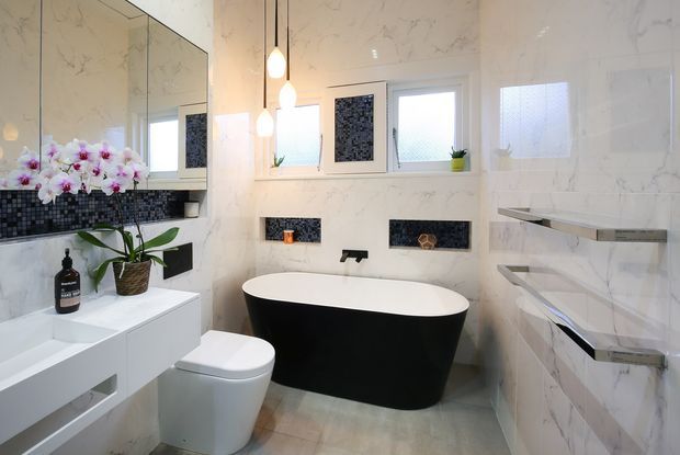 Contemporary bathroom featuring a freestanding black-and-white bathtub, wall-mounted black tapware, wall-hung vanity, toilet, mirrored cabinetry, and open glass shelving. Large white marble-look wall tiles are paired with small black mosaic tile feature niches and a light neutral floor. Frosted windows and pendant lights provide soft, bright illumination, while orchids add a decorative touch.