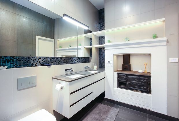 A contemporary bathroom featuring a floating white double vanity with integrated basins, large mirrored wall cabinetry, dark tiled flooring, pale wall tiles, and a blue-grey mosaic splashback. Open white shelving with integrated lighting surrounds a traditional cast-iron wood stove set into a white-painted brick fireplace surround. The room has bright task lighting and a refined black-and-white palette with blue mosaic accents.