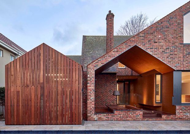 Street-facing view of a compact contemporary brick residence with a sculptural projecting entry canopy, warm timber soffit and recessed timber entry door. The facade combines red brick masonry, dark metal framing, vertical timber screening and a large timber garage or gate structure, with pitched roof forms and a prominent brick chimney.