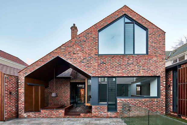 Exterior view of a contemporary two-storey brick residence in a dense suburban setting. The home features a steep front gable, extensive red brickwork, a large upper-level glazed gable window, dark-framed windows, and a recessed covered entry or courtyard with timber-lined surfaces. Adjacent homes and a paved forecourt are visible.
