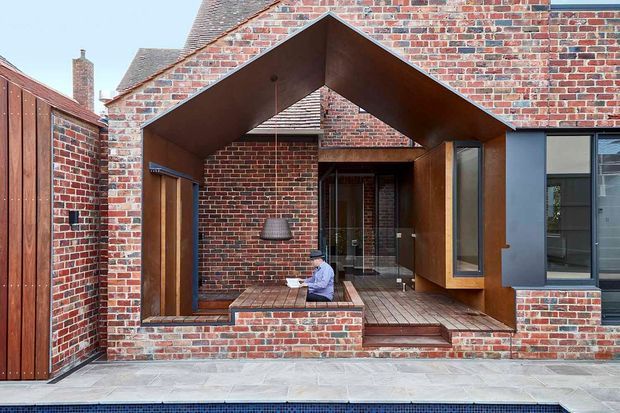 Contemporary brick house facade with a dramatic projecting, angular entrance canopy clad in dark metal or timber-toned panels. The composition includes exposed red brick walls, dark-framed glazing, timber decking, a paved forecourt, and a small recessed entry courtyard. A pool edge is visible along the bottom of the image.