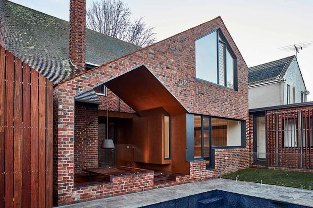 Exterior view of a contemporary brick residential extension beside a swimming pool. The design combines red exposed brick walls, dark metal-framed glazing, a projecting angular timber-lined volume, and a sheltered courtyard-like outdoor space. Existing pitched-roof brick and weatherboard buildings are visible around the extension.