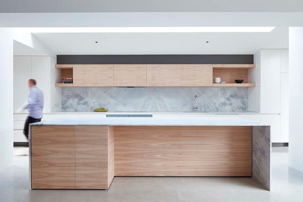 A bright, contemporary open-plan kitchen featuring a large wood-clad island with a white benchtop, pale timber overhead cabinetry, integrated white side cabinetry, and a dramatic veined stone splashback extending across the rear wall and island end panel. The space has a clean white ceiling and walls, recessed lighting, a polished light floor, and strong horizontal built-in joinery. A blurred person is visible at the side.