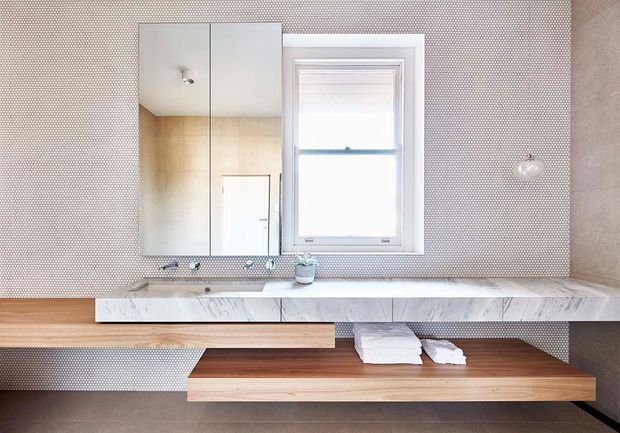 Bright contemporary bathroom featuring a long floating marble vanity with integrated twin basins, wall-mounted taps, a timber open shelf beneath, mirrored cabinetry, a white-framed window, and small-format patterned wall tiles. A freestanding glass vessel and folded towels add minimal styling.