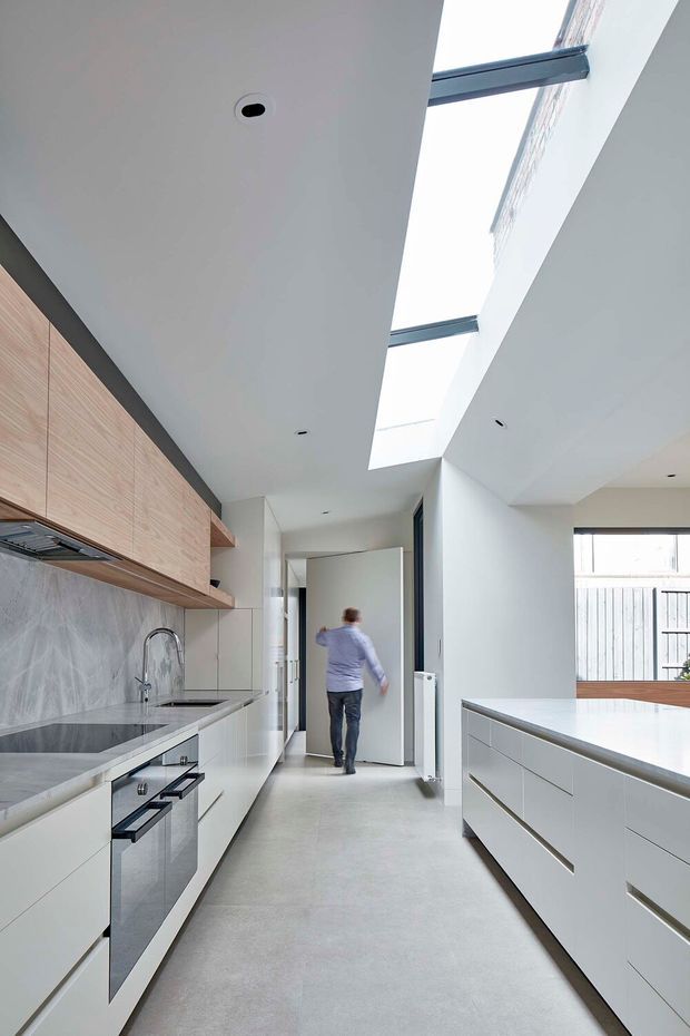 Bright contemporary kitchen viewed along a galley-style circulation space, with extensive white flat-panel cabinetry, pale timber overhead cupboards, a grey stone-look splashback, integrated appliances, and a large central island. A long skylight brings strong natural light into the room, while a glazed opening at the rear connects visually to an exterior or adjacent space.
