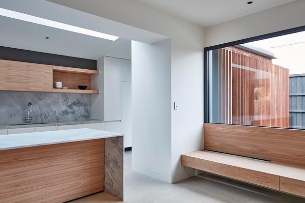 A bright, contemporary open-plan kitchen and adjoining window seat. The kitchen features warm timber cabinetry and open shelving, a pale stone island benchtop, a dramatic grey stone splashback, and integrated white cabinetry. A built-in timber window bench with drawers sits beside large black-framed glazing, looking toward a timber-clad exterior screen. The room has a clean, minimalist composition with recessed downlights and a light neutral palette.