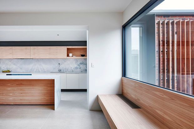 Bright contemporary open-plan kitchen and window-seat area. The kitchen features light timber cabinetry and island panels, a pale marble-look splashback, white walls and cabinetry, and a smooth light-grey floor. A built-in timber window seat runs along expansive dark-framed glazing, with vertical timber screening visible outside. The composition is clean-lined and minimalist with abundant natural light.