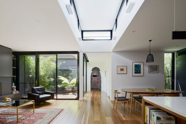 Bright open-plan contemporary living and dining area with warm timber flooring, extensive black-framed glazing, a central skylight, and a white sculptural ceiling volume. The living zone includes a dark leather armchair, patterned rug and low coffee table, while the dining area has a timber table, pendant light and built-in shelving. An arched hallway opening leads toward the rear of the home.