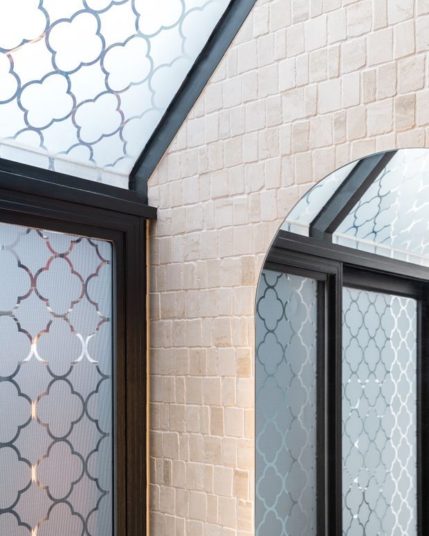 Close-up architectural detail of a pale exposed brick wall with black-framed arched and angled glazing. The glass features a repeating decorative quatrefoil pattern, creating a refined contrast between the light masonry and dark metal framing.