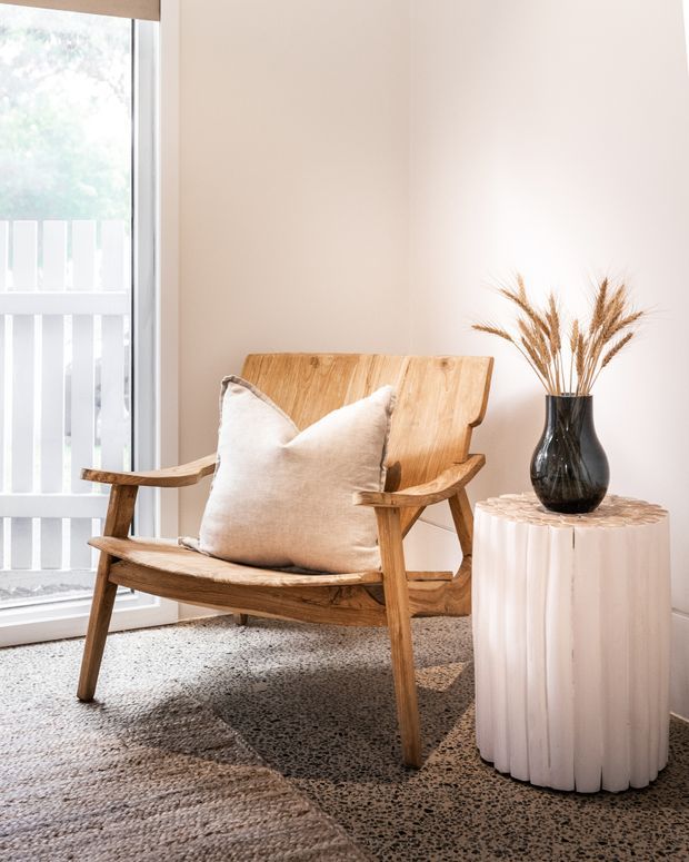 A bright, softly styled corner featuring a sculptural timber lounge chair with a cream cushion beside a fluted white pedestal table topped with a dark glass vase and dried grasses. A large glazed opening with white vertical balustrade elements brings in abundant natural light. The space has pale warm walls and a textured neutral carpet layered over a speckled hard floor.