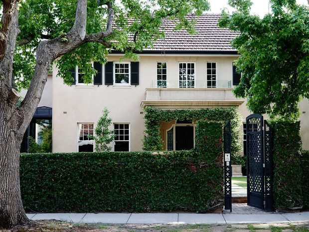 Exterior view of a refined two-storey traditional residence, partially screened by mature trees and a clipped hedge. The pale rendered facade has symmetrical multi-pane windows with dark shutters, a tiled pitched roof, a projecting classical-style entrance canopy, and an ornate dark metal pedestrian gate opening onto a paved path. Climbing greenery surrounds the entry.