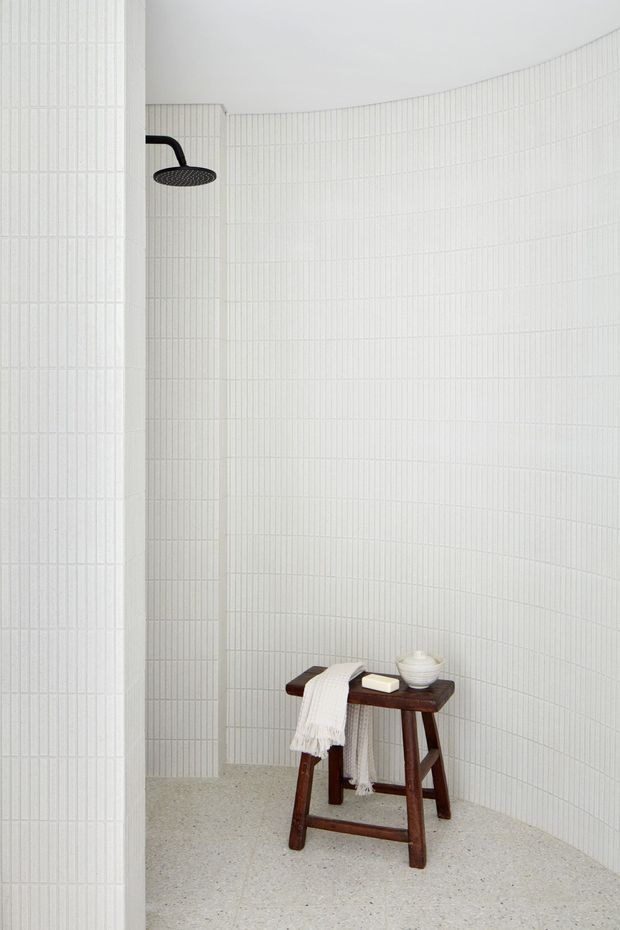 Minimalist walk-in shower space with curved walls fully clad in fine white linear tiles. A matte black round showerhead contrasts with the white surfaces, while a dark timber stool holding folded towels and a small ceramic vessel adds warmth. The floor is a light speckled cementitious or terrazzo-like finish, and the space is bright, clean, and serene.