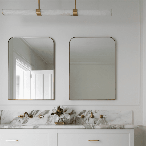 A refined double vanity bathroom featuring white cabinetry, a dramatic white-and-grey veined marble backsplash and countertop, brass tapware and hardware, twin rounded-corner mirrors, and a slim brass-and-glass wall light. The palette is light, warm, and elegant with a classic contemporary feel.