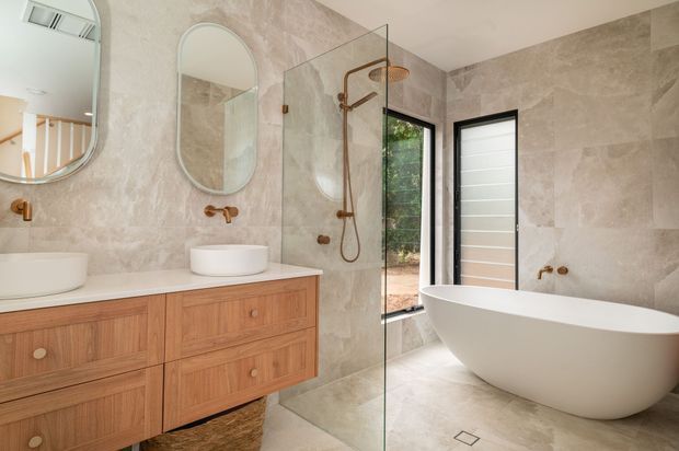 A bright contemporary bathroom with large beige stone-look wall and floor tiles, a warm timber floating vanity with two white vessel basins, arched mirrors, brushed copper tapware, a glass-enclosed shower, and a sculptural freestanding white bathtub. Black-framed glazing and a timber garden view add contrast and natural light.