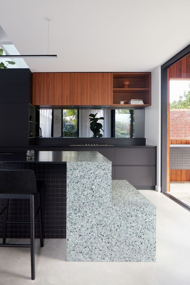 A contemporary kitchen with a dark, refined palette and a prominent pale terrazzo island with stepped seating. The kitchen features matte dark cabinetry, a black tiled backsplash, warm walnut overhead joinery with open shelving, stainless-steel appliances and fittings, and black-framed glazing opening to an outdoor brick-and-timber area. A linear pendant light and indoor planting add understated detail.