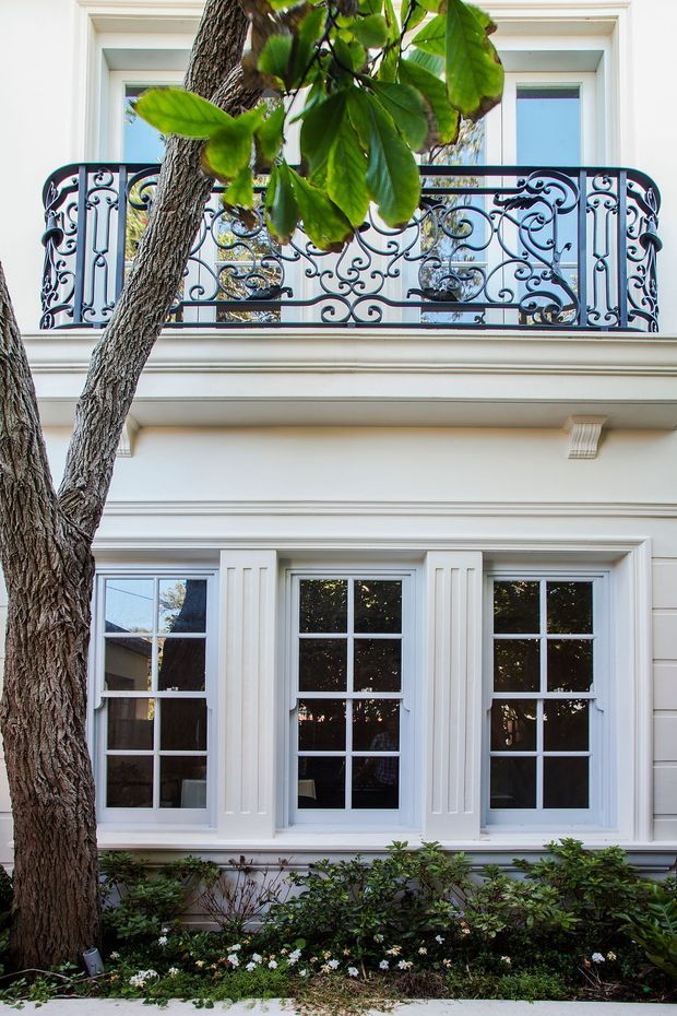 Exterior view of a refined multi-storey facade with white painted detailing, tall multi-pane windows, an ornate black wrought-iron balcony balustrade, and classical mouldings. Mature tree branches and landscaped planting partially frame the building.