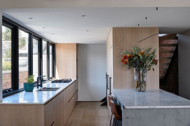 Bright contemporary kitchen with a long galley-style run of timber cabinetry beneath black-framed windows, a pale stone benchtop, integrated gas cooktop and sink, and a large central island with a grey stone surface and concrete-look base. A timber-panelled volume conceals storage and separates the kitchen from an exposed brick stairway, while a vase of flowers adds colour. The space has recessed ceiling lights, warm timber flooring, and abundant natural light.