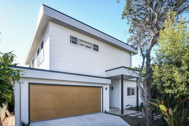 Exterior view of a contemporary two-storey residence with clean white horizontal cladding, a low-pitched sloping roof, dark trim, and a broad timber-toned garage door. A covered entry sits beside the garage, with landscaped planting and mature trees framing the house.