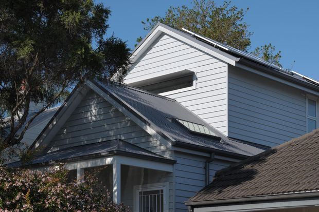 Exterior view of a light grey weatherboard house with intersecting pitched gable roofs, corrugated metal roofing, white trim, and a small upper-level horizontal window. Mature trees and a neighbouring roof partially frame the building.