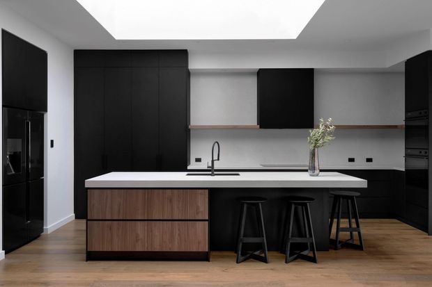 Contemporary kitchen with matte black full-height cabinetry, a large white island with integrated sink and dark bar stools, warm timber island cabinetry and flooring, and a large ceiling skylight. Built-in black appliances and open shelving complete the streamlined design.