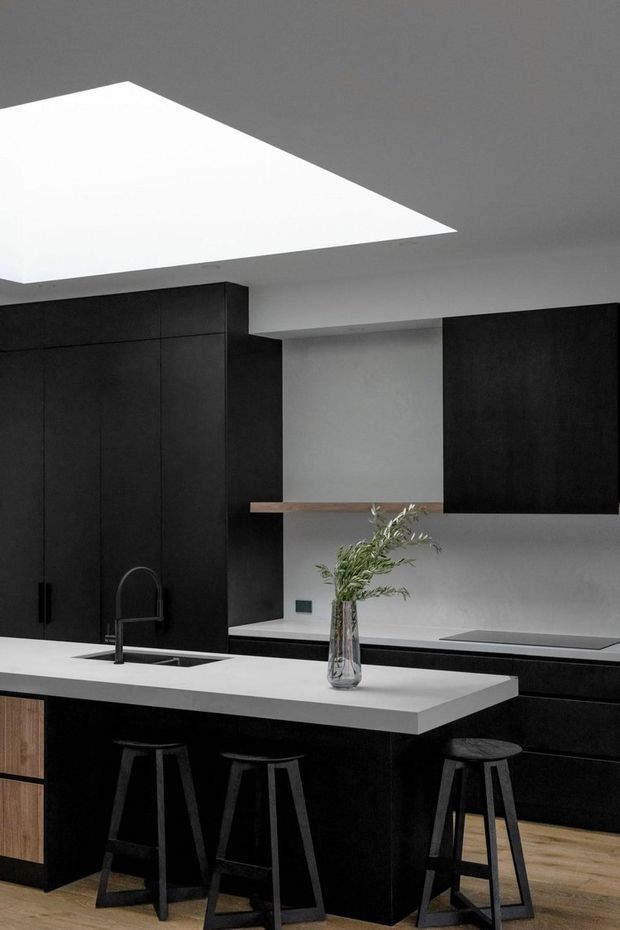 A contemporary black-and-white kitchen with matte-black full-height cabinetry, a white island benchtop with integrated sink and dark stools, pale timber flooring, and a simple timber shelf against a light wall. A large geometric ceiling opening or skylight provides strong overhead illumination, while a glass vase with greenery adds a small natural accent.