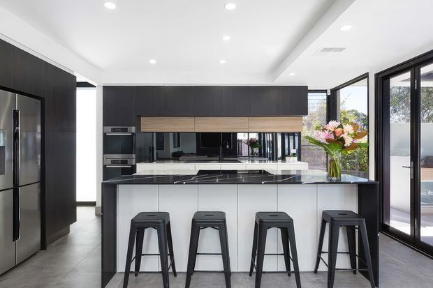 A bright contemporary kitchen with a large central island and four black bar stools. The space features dark charcoal cabinetry, horizontal timber-toned overhead joinery, white cabinetry and benchtops, a dark stone island benchtop, integrated appliances, recessed ceiling lights, and expansive black-framed glazing opening to the outdoors.
