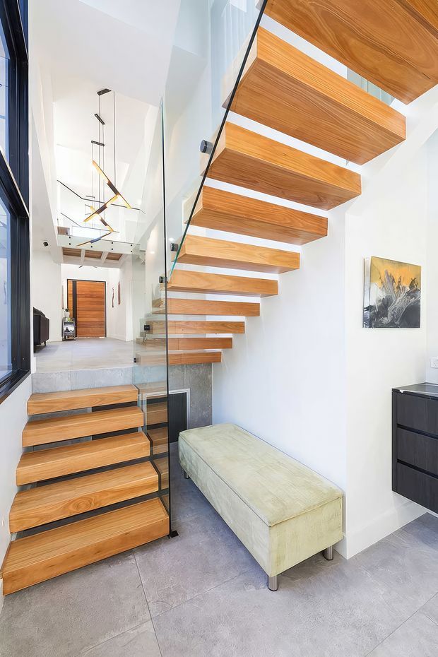 A bright contemporary stair hall featuring a sculptural open-riser staircase with thick warm timber treads and matching floating upper steps. A clear glass balustrade with dark metal fittings runs alongside the stairs. White walls, large grey-format floor tiles, a pale upholstered bench, dark built-in cabinetry, modern pendant lighting, and a timber entry door complete the space.