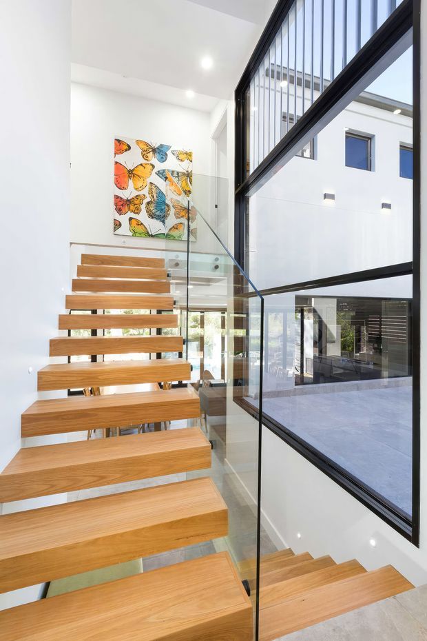 Bright contemporary stairway with floating light-wood treads, clear glass balustrades, and dark-framed floor-to-ceiling glazing overlooking an internal courtyard or adjacent space. White walls and ceilings create an airy, minimalist setting, with a colourful butterfly artwork on the upper landing.