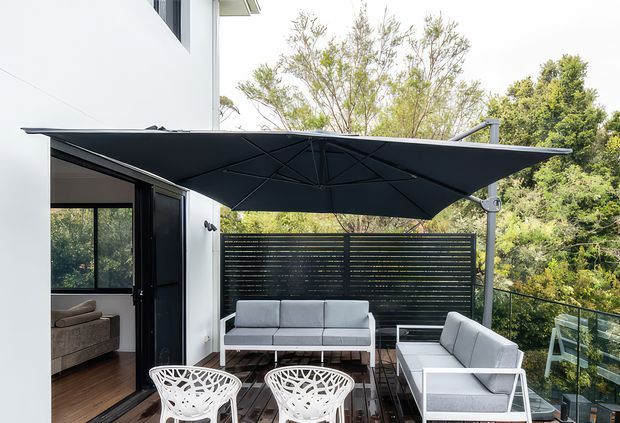 Contemporary outdoor terrace adjoining a white modern building, furnished with a grey outdoor sofa, armchairs and white sculptural chairs beneath a large dark cantilevered shade canopy. A horizontal slatted privacy screen, glass balustrade and surrounding trees frame the elevated deck, with a glimpse of the interior living room through the open doors.