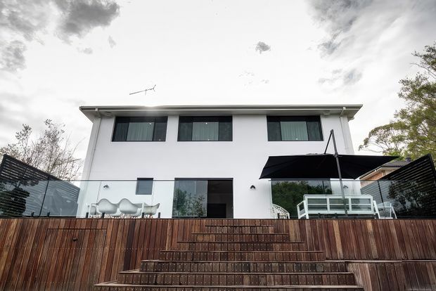 Low-angle exterior photograph of a contemporary two-storey white residence with a flat, slightly projecting roof, dark-framed rectangular windows, and large glazed openings. A timber-clad stepped terrace or deck occupies the foreground, with glass balustrades, outdoor seating, and a large black patio umbrella. Trees and an overcast sky surround the property.