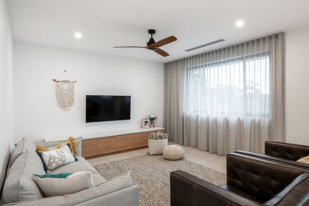 Bright contemporary living room with a light neutral palette, white walls, a timber media console, wall-mounted television, large curtained window, ceiling fan, textured rug, light sofa, and dark leather armchairs.