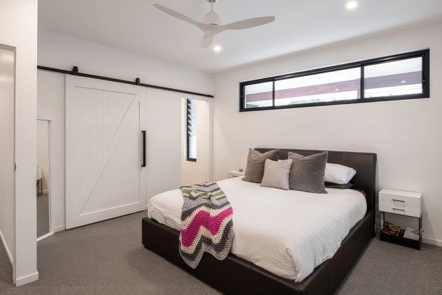 A bright, contemporary bedroom with a large dark upholstered bed, white bedding, layered neutral cushions, and a colourful knitted throw. White walls and a ceiling fan create an airy feel, while a wide black-framed clerestory-style window provides natural light. A white sliding barn-style wardrobe door with black hardware forms a strong feature wall, and grey carpet covers the floor.