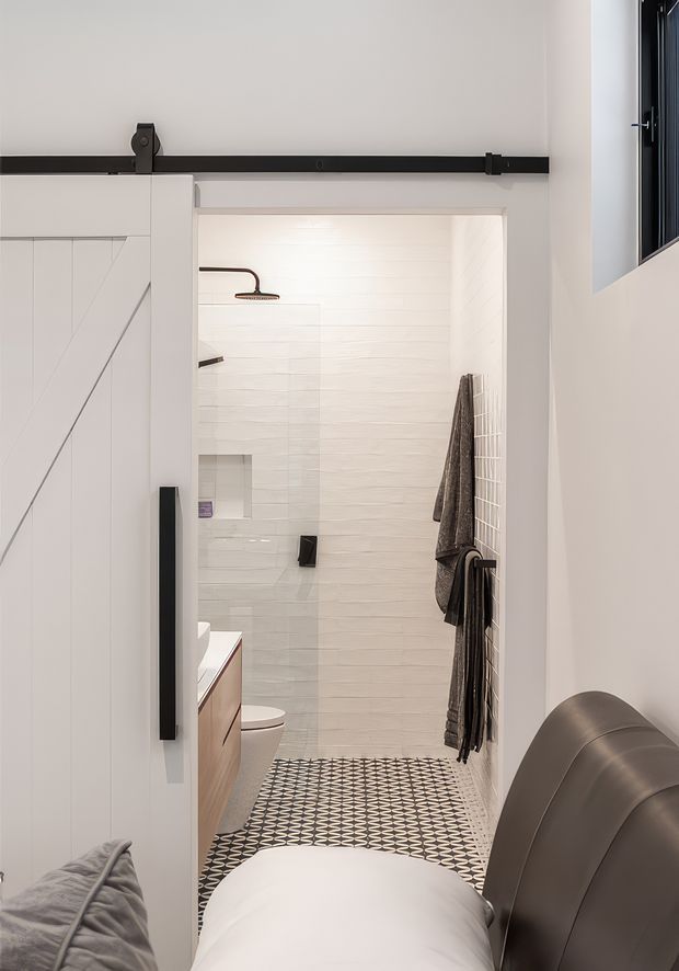 A compact modern bathroom viewed from an adjacent bedroom through a partially open white barn-style sliding door. The bathroom features white horizontally textured wall tiles, a glass shower enclosure with dark fixtures, a timber vanity, toilet, patterned black-and-white floor tiles, and dark towels on a wall-mounted rail. A portion of the bedroom bed is visible in the foreground, clearly showing the bathroom connection.