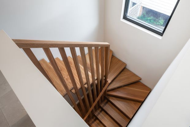 View looking down into a compact modern stairwell with warm timber stair treads, matching timber balustrade and vertical spindles, white walls, and a high-set black-framed window providing natural light.