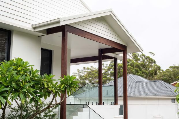 Exterior view of a contemporary home extension or covered entry terrace, featuring white horizontal weatherboard cladding, a pitched gable roof, dark timber posts and beams, and clear glass balustrading. The elevated terrace overlooks neighbouring rooftops and greenery.