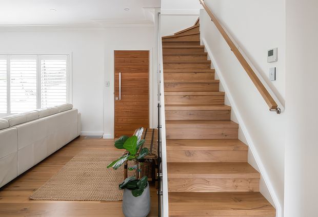 Bright contemporary entryway showing a timber staircase with open risers or minimal riser detailing, a matching timber handrail, white walls, and timber flooring. A large timber front door, woven rug, potted plants, and part of an adjacent living room with a pale sofa are visible. White plantation-style shutters provide filtered daylight.