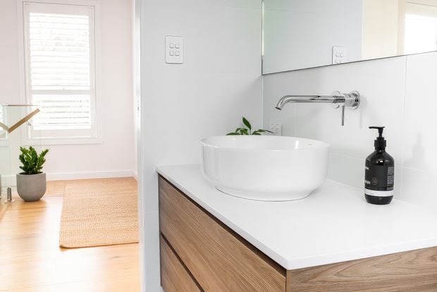 Bright contemporary bathroom vanity with a white vessel basin, wall-mounted chrome tapware, white tiled walls, and a timber-look floating vanity with a white benchtop. A large mirror reflects the room, while a window, plants, and a woven mat add natural accents.