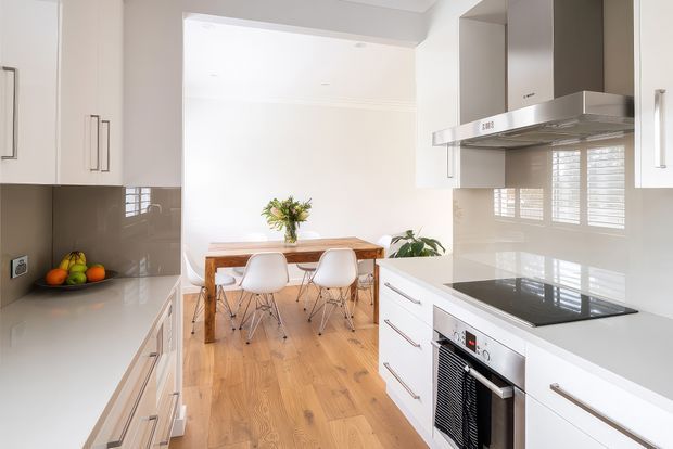 Bright contemporary white kitchen opening directly into a dining area. The kitchen features white flat-panel cabinetry, a pale stone-look benchtop and splashback, a black glass induction cooktop, stainless-steel rangehood and integrated oven. A timber dining table with white chairs sits beyond the kitchen, over warm timber flooring, with white plantation shutters and abundant natural light.