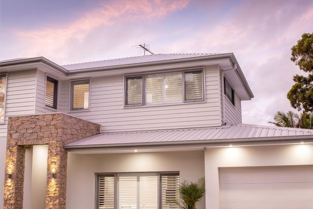 Twilight exterior view of a two-storey suburban house with pale horizontal weatherboard cladding, a light metal hipped roof, broad eaves, horizontal louvre-style windows, and a prominent rough stone-clad entry feature. A garage and covered ground-floor area are visible.