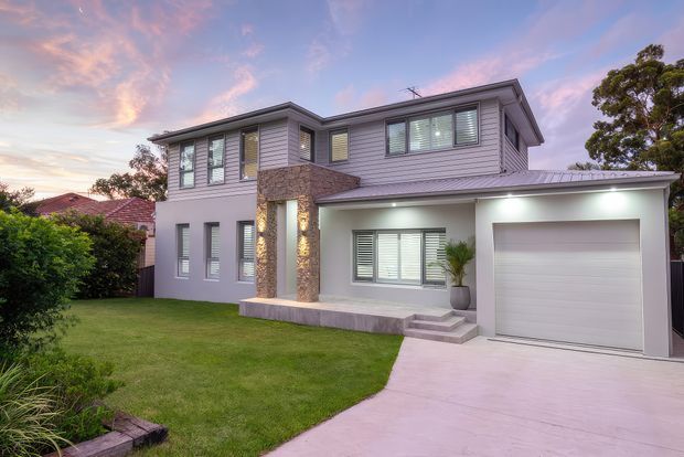 A contemporary two-storey suburban house photographed at dusk. The facade combines light grey horizontal cladding, rendered walls, and a textured stone-clad entry feature with warm integrated lighting. A single garage, upper-level windows with external shutters, front lawn, and pale concrete driveway are visible.