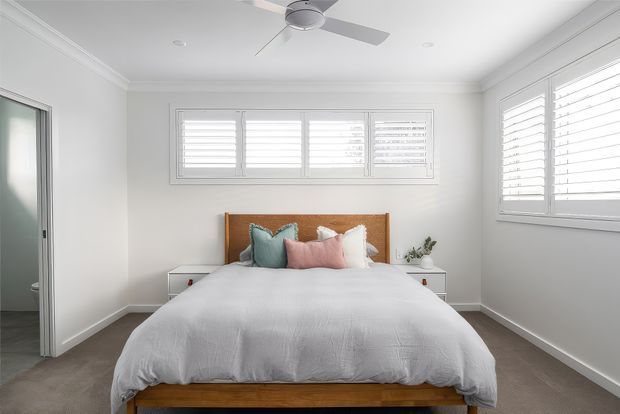 Bright, clean bedroom with a low-profile timber bed and warm timber headboard, layered white bedding, pastel blue and pink cushions, white walls and plantation shutters. A ceiling fan, built-in-style bedside tables, and ample shuttered windows create an airy contemporary coastal feel.